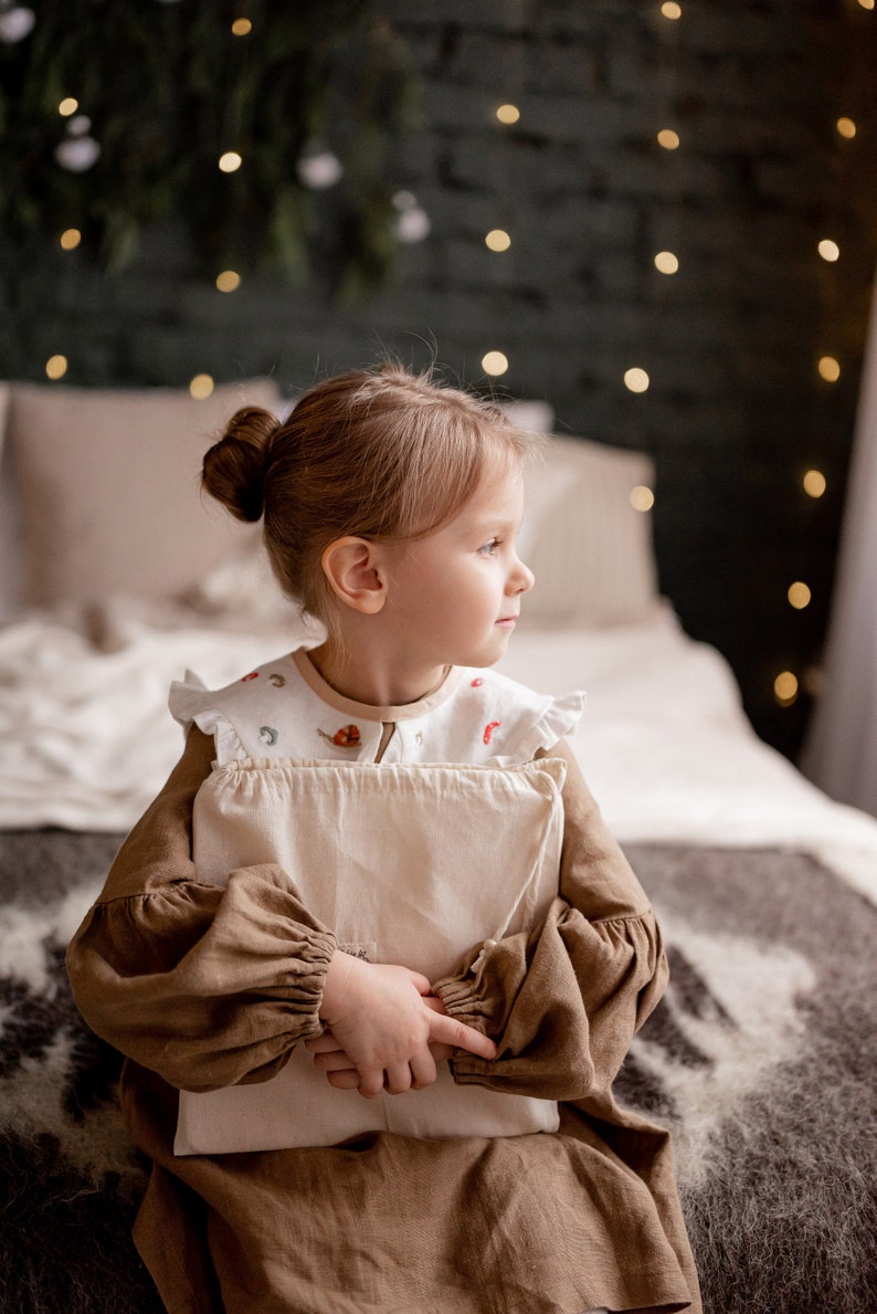 Op de afbeelding: Een jong meisje in een bruine jurk zit op een bed en houdt een wit kussen vast. Ze kijkt naar de zijkant en de achtergrond is wazig.