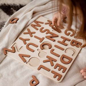Op de afbeelding: Een kind speelt met een houten alfabetpuzzel. De puzzel heeft alle letters van het alfabet in bruine houten stukjes. Het kind plaatst de letter "C" in zijn gleuf op het bord.