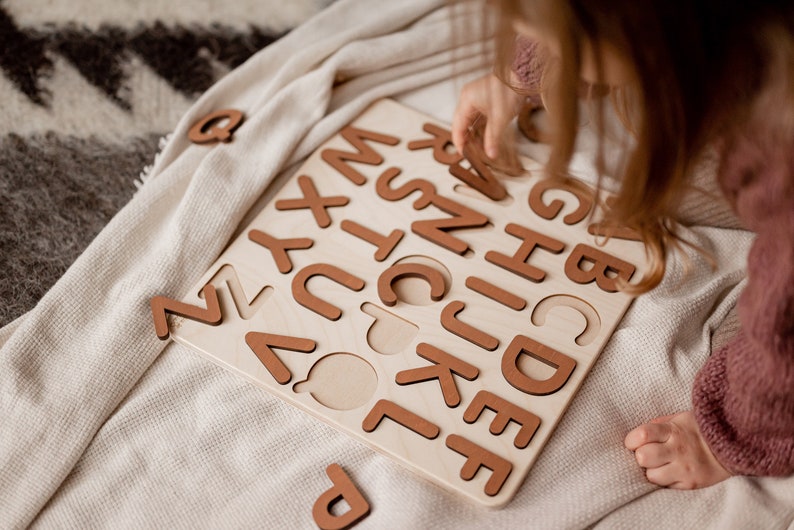 Op de afbeelding: Een kind speelt met een houten alfabetpuzzel. De puzzel heeft alle letters van het alfabet in bruine houten stukjes. Het kind plaatst de letter "C" in zijn gleuf op het bord.