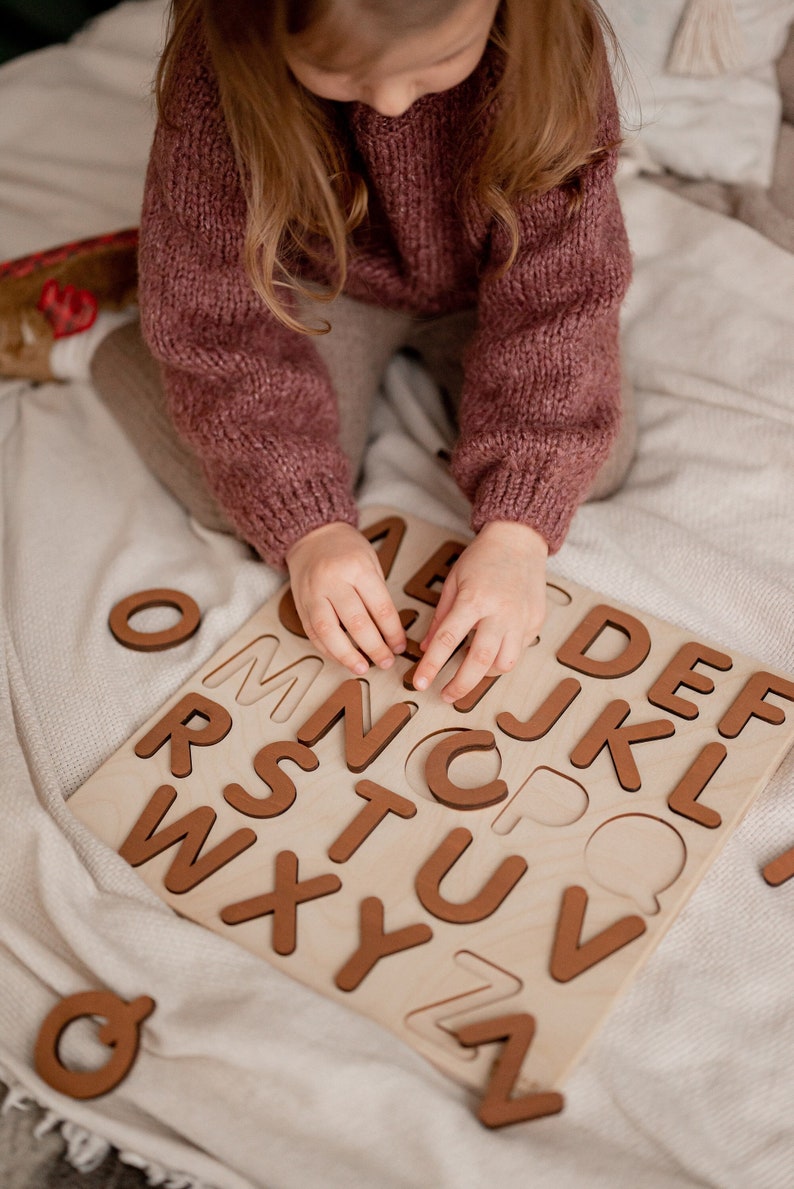Op de afbeelding: Een kind speelt met een houten alfabetpuzzel. De puzzel heeft alle letters van het alfabet in afzonderlijke stukken. Het kind houdt de letter C in zijn hand.