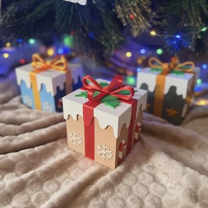 Könnte beinhalten: Drei quadratische Geschenkboxen mit Deckeln, die weiße, wellenförmige Ränder wie Schnee aufweisen. Die Boxen sind in Rot, Blau und Dunkelgrau gehalten, mit weißen Schneeflocken, Zuckerstangenmustern und roten oder orangen Schleifen.
