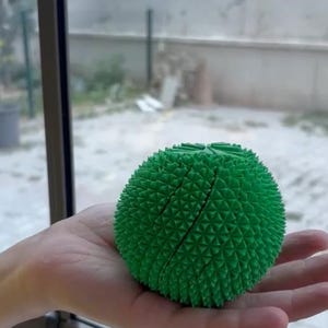 May include: A bright green spherical object with a textured, geometric surface composed of numerous small, pointed triangular pyramids. The object features a distinct vertical seam running across its centre, dividing the textured surface.