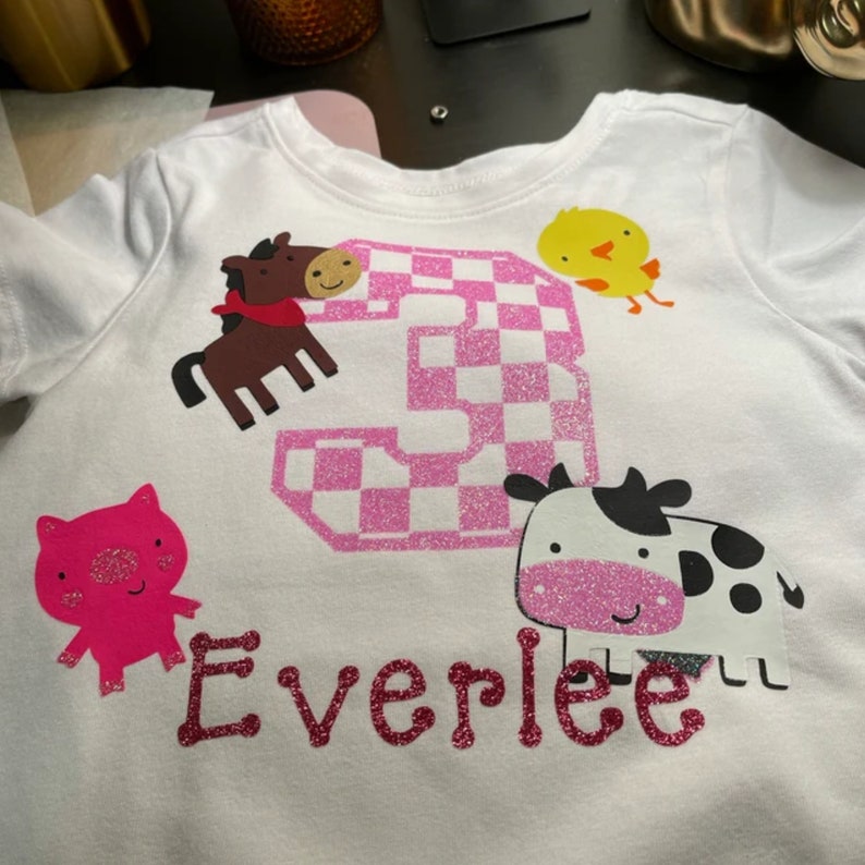 K&ouml;nnte beinhalten: Ein wei&szlig;es T-Shirt mit einer pinkfarbenen glitzernden Zahl drei, einem rosa Schwein, einem braunen Pferd, einem gelben K&uuml;ken, einer schwarz-wei&szlig;en Kuh und dem Namen Everlee in pinkfarbenem Glitzer.
