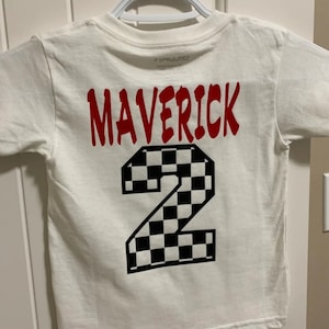K&ouml;nnte beinhalten: Wei&szlig;es T-Shirt mit rotem Text "Maverick" und einer schwarz-wei&szlig;en karierten Nummer "2" auf der R&uuml;ckseite.