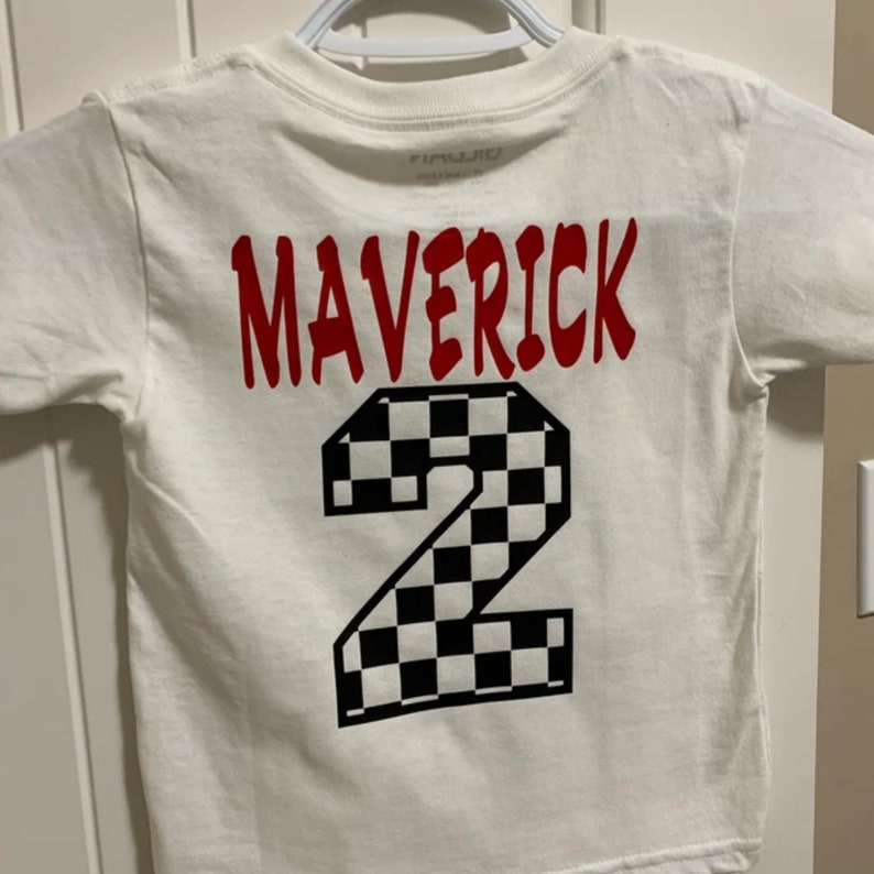 K&ouml;nnte beinhalten: Wei&szlig;es T-Shirt mit rotem Text "Maverick" und einer schwarz-wei&szlig;en karierten Nummer "2" auf der R&uuml;ckseite.