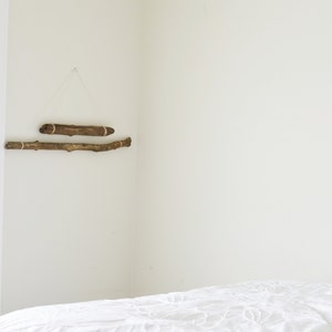May include: Two pieces of driftwood are hung on a white wall with twine. The driftwood is arranged horizontally, one above the other. The driftwood is a natural brown color. A white bed with a white comforter is in the foreground.