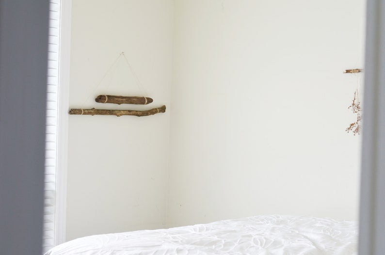 May include: Two pieces of driftwood are hung on a white wall with twine. The driftwood is arranged horizontally, one above the other. The driftwood is a natural brown color. A white bed with a white comforter is in the foreground.