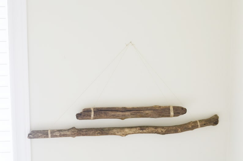 May include: Two pieces of driftwood are hung on a white wall with twine. The driftwood is brown and has a natural, rustic look.