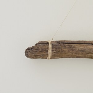 May include: A piece of driftwood with a natural, weathered finish is tied with twine and hangs from a white wall.