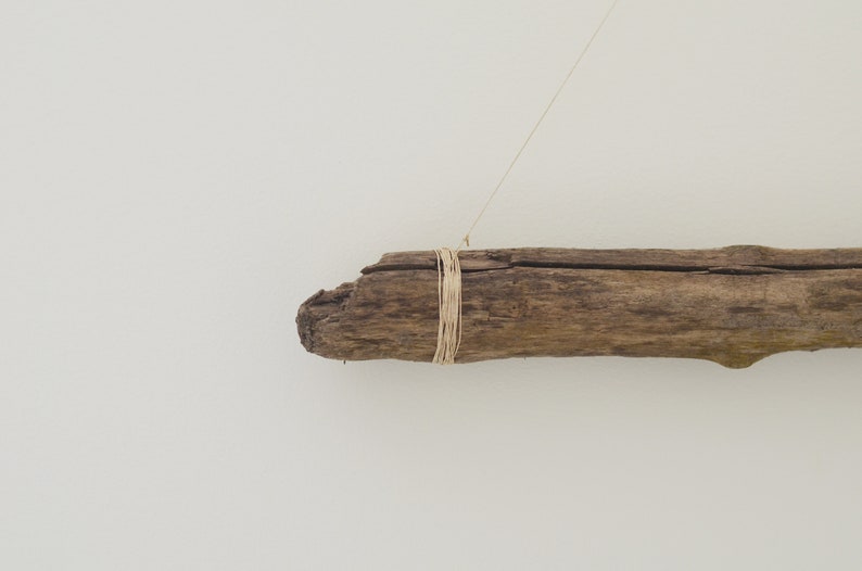 May include: A piece of driftwood with a natural, weathered finish is tied with twine and hangs from a white wall.