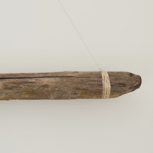 May include: A piece of driftwood with a natural brown color is hanging from a string. The driftwood is tied with a thin, natural-colored twine.