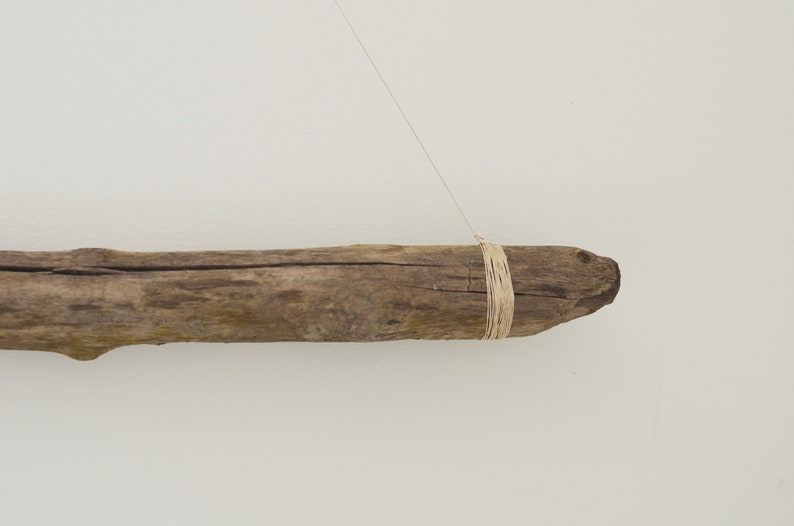 May include: A piece of driftwood with a natural brown color is hanging from a string. The driftwood is tied with a thin, natural-colored twine.