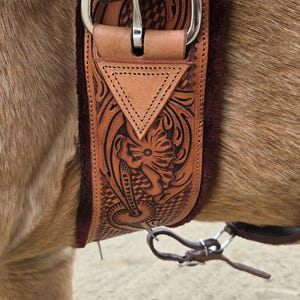 May include: Close-up of a brown leather horse saddle with intricate floral and geometric carvings. The saddle features a silver buckle and a soft, burgundy-colored underside. The saddle is attached to a horse.