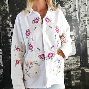 Può includere: Una camicia bianca a maniche lunghe con bottoni, caratterizzata da ricami floreali colorati e dettagli in pizzo sul davanti e sulle maniche. La camicia include due tasche frontali con motivi floreali e un colletto, abbinata a una bandana con margherite.