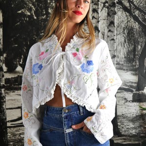 May include: White embroidered bolero jacket with floral designs in pink, blue, and yellow. The jacket has long sleeves and a tie closure. The model is wearing a blue denim skirt and a navy blue bandana.