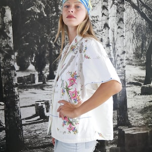 Può includere: Una camicia bianca a maniche corte con bottoni, ricami floreali colorati e un foulard con stampa di limoni blu e gialli. La camicia presenta un colletto e una vestibilità ampia, abbinata a pantaloncini di jeans azzurri con orlo sfrangiato.
