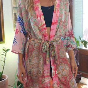 Peut inclure: Un peignoir kimono à manches longues présentant un mélange de motifs roses, rouges et sarcelle, incluant des imprimés cachemire et floraux. Le vêtement possède une ceinture à nouer et une bande verticale rose tie-dye sur le devant.
