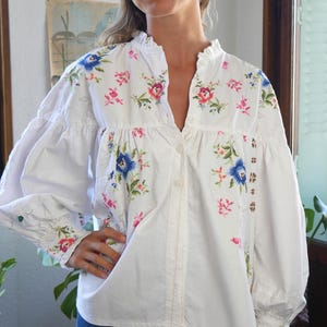 May include: A white long-sleeved button-down blouse featuring a ruffled collar and a colorful floral pattern with blue, pink, and green embroidered details. The garment has voluminous sleeves with buttoned cuffs and a loose, gathered fit.