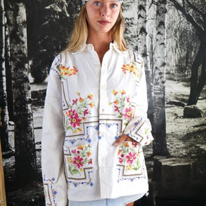 Può includere: Una camicia a maniche lunghe con bottoni color bianco sporco caratterizzata da un colorato ricamo floreale con accenti rosa, gialli e verdi disposti in cornici geometriche, abbinata a un foulard bianco e nero con motivo a margherite.
