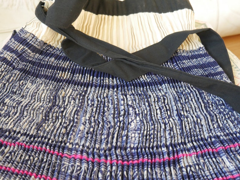 May include: A pleated skirt with a blue and white patterned design. The skirt has a black ribbon tied around the waist and a pink stripe at the bottom.