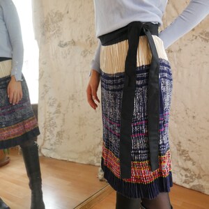 May include: A pleated skirt with a black ribbon belt. The skirt is made of a blue and white patterned fabric with a colorful embroidered trim.