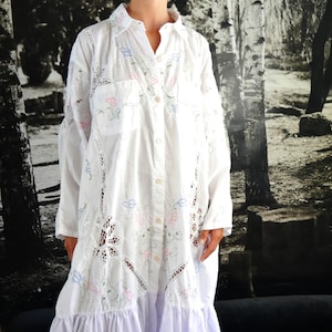 Könnte beinhalten: Ein langärmeliges, weißes Hemdblusenkleid mit aufwendigen Lochstickereien und zarten Blumenmustern in Pastellrosa, Blau und Grün. Das Kleidungsstück verfügt über einen Kragen, eine Brusttasche und einen breiten, gerafften Volantsaum.