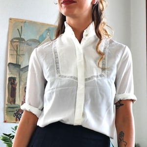 Peut inclure: Chemisier blanc à col montant et devant plissé. Le chemisier présente des détails en dentelle autour du col et une fermeture boutonnée. Les manches sont retroussées. Il est associé à une jupe plissée bleu foncé.