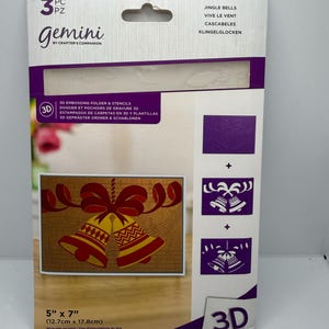 May include: A 3-piece Gemini by Crafter's Companion 3D embossing folder and stencil set titled Jingle Bells. The packaging displays a 12.7 cm by 17.8 cm finished card design featuring two gold and red bells with a red bow on a musical note background.