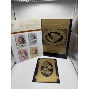 May include: A gold metal craft die set for card making, including an instruction sheet labeled Shadow Box Card Die Set by Anna Griffin. The set features an ornate oval frame with floral cutouts and a separate rectangular die with floral motifs.