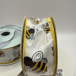 Op de afbeelding: Een rol wit lint met een herhaald patroon van zwarte en gele bijen en krullen. Het lint heeft een gele gestikte rand. De rol zit op een witte kartonnen spoel. De tekst "32 YDS ESTER" is zichtbaar op de spoel.