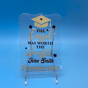 Può includere: Un cartello in acrilico trasparente con un cappello di laurea con glitter dorati e il testo "The tassel was worth the hassle" in nero. Il nome "John Smith" e "Graduation 2024" sono anch'essi in nero.
