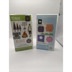 May include: Two rectangular Cricut cartridges in plastic cases. The left case is green and white, labelled Outdoor Man, featuring graphics of trees, a campfire, and a person. The right case is blue and white, labelled Elegant Edges, showing various die-cut shapes.