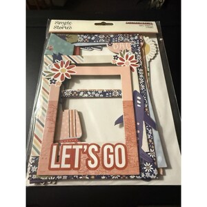 May include: A package of Simple Stories Chipboard Frames with travel-themed designs. The frames feature floral accents, a suitcase, and a purple airplane. The phrase "LET'S GO" is prominently displayed in red letters.
