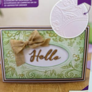 Puede incluir: Un paquete de carpeta de relieve 3D que muestra una tarjeta verde con patrón floral, un lazo de arpillera color canela y la palabra Hello en cursiva. El empaque muestra las dimensiones de 15,2 cm por 10,2 cm y destaca un efecto 3D.
