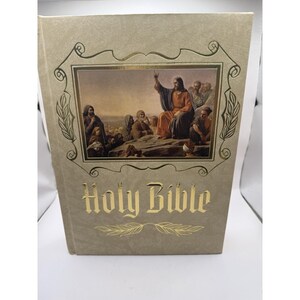 May include: A hardcover Holy Bible with a textured beige cover featuring a gold-foiled rectangular frame surrounding a printed illustration of a figure teaching a group of people. The words Holy Bible are printed in gold lettering below the image.