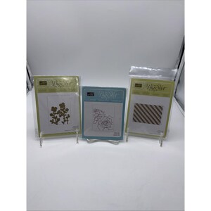 May include: Three Big Shot embossing folders in clear plastic packaging. The folders feature floral and geometric designs. One folder has a floral design, another has a rose, and the third has diagonal stripes. The folders are displayed on clear stands.