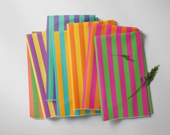 May include: A stack of colorful striped paper sheets in shades of purple, yellow, orange, teal, and pink. A sprig of greenery rests on top of the pink and green striped sheet. The sheets are arranged on a white surface.
