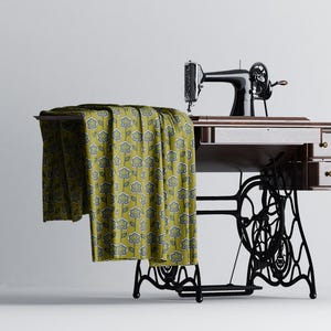 以下が含まれることがあります： 黒い金属製のフレームと木製の天板を持つヴィンテージのミシン。テーブルの上に花柄の黄色の布がかけられています。