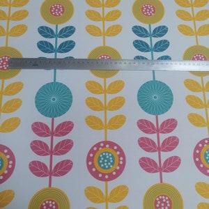 May include: White fabric with a repeating floral pattern. The design features stylized flowers in yellow, pink, and teal, with yellow leaves and teal stems. A metal ruler is placed across the fabric.
