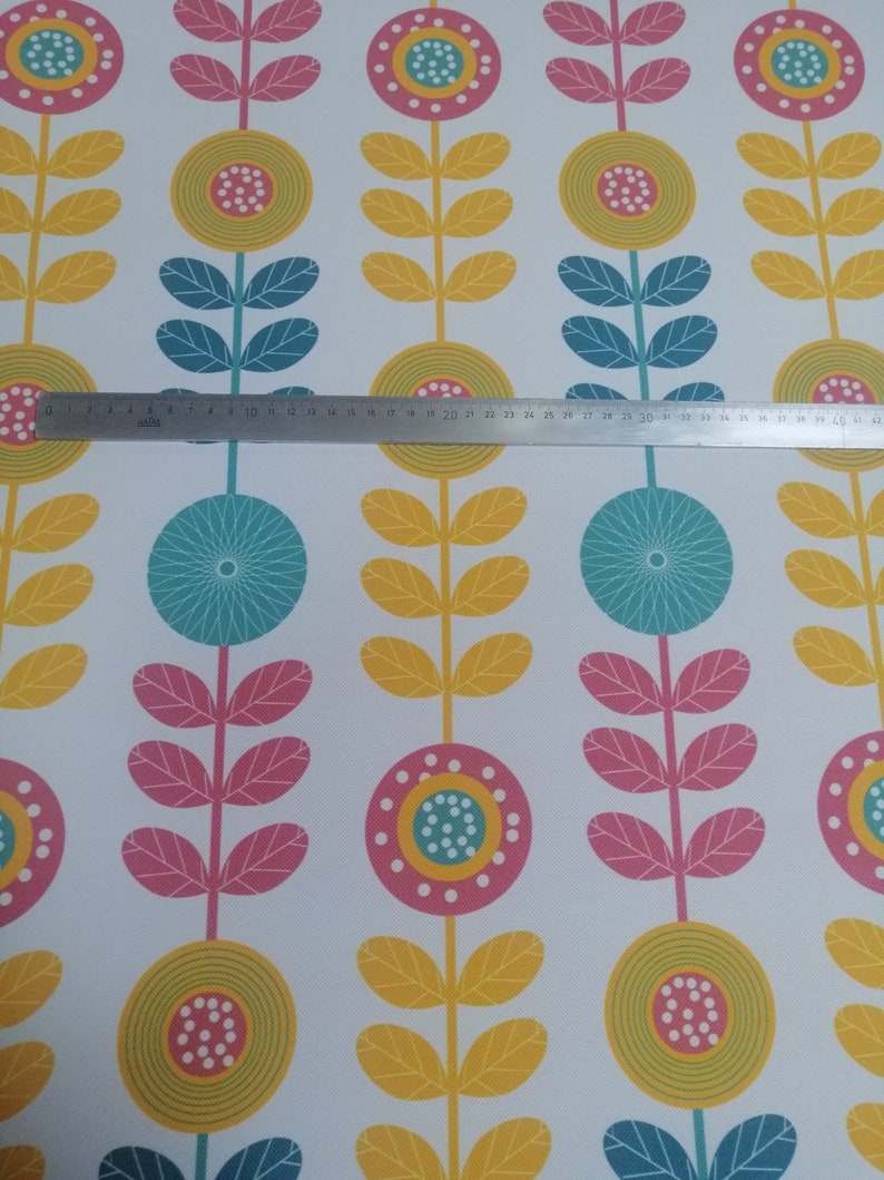May include: White fabric with a repeating floral pattern. The design features stylized flowers in yellow, pink, and teal, with yellow leaves and teal stems. A metal ruler is placed across the fabric.