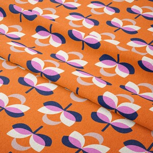 K&ouml;nnte beinhalten: Nahaufnahme eines orangefarbenen Stoffes mit einem sich wiederholenden Blumenmuster. Die Blumen bestehen aus wei&szlig;en, rosa und marineblauen Bl&uuml;tenbl&auml;ttern mit einem hellgrauen, gebogenen Akzent. Der Stoff hat ein strukturiertes Aussehen.