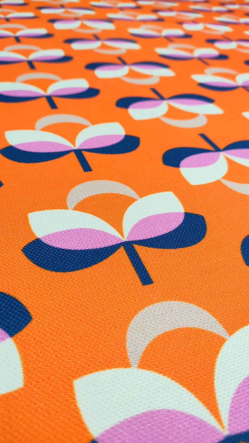 K&ouml;nnte beinhalten: Nahaufnahme eines Stoffes mit einem sich wiederholenden Blumenmuster. Die Blumen haben wei&szlig;e, rosa und beige Bl&uuml;tenbl&auml;tter mit marineblauen und schwarzen Stielen und Akzenten, vor einem leuchtend orangefarbenen Hintergrund. Der Stoff hat ein strukturiertes Aussehen.