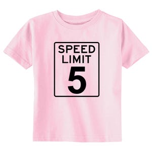 Pode incluir: Uma t-shirt de manga curta em rosa claro com uma impress&atilde;o gr&aacute;fica preta de um sinal retangular de limite de velocidade. O sinal cont&eacute;m o texto SPEED LIMIT em letras mai&uacute;sculas pretas acima de um grande n&uacute;mero 5 preto.