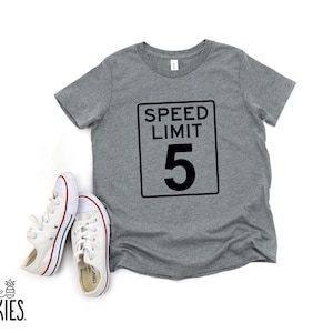 Pode incluir: Uma t-shirt cinzenta mesclada de manga curta com um gr&aacute;fico preto de um sinal retangular de limite de velocidade com o texto SPEED LIMIT 5. Ao lado da t-shirt est&aacute; um par de t&eacute;nis de lona brancos com riscas vermelhas e azuis nas solas.