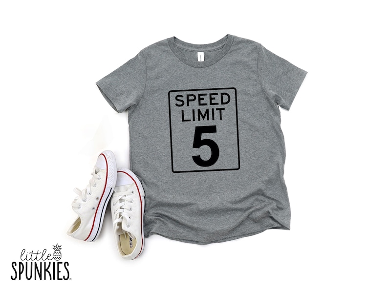 Pode incluir: Uma t-shirt cinzenta mesclada de manga curta com um gr&aacute;fico preto de um sinal retangular de limite de velocidade com o texto SPEED LIMIT 5. Ao lado da t-shirt est&aacute; um par de t&eacute;nis de lona brancos com riscas vermelhas e azuis nas solas.