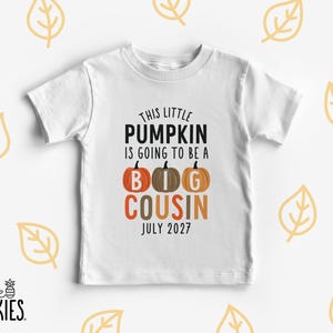 May include: A white short-sleeved t-shirt featuring the black and orange text THIS LITTLE PUMPKIN IS GOING TO BE A BIG COUSIN JULY 2027. The design includes three illustrated pumpkins in orange and brown colors with white letters spelling BIG.