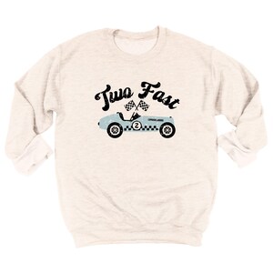 May include: A cream-colored long-sleeve sweatshirt featuring a distressed black graphic that reads Two Fast above a light blue vintage race car with a checkered flag design, the number two on the side, and two crossed checkered flags above it.