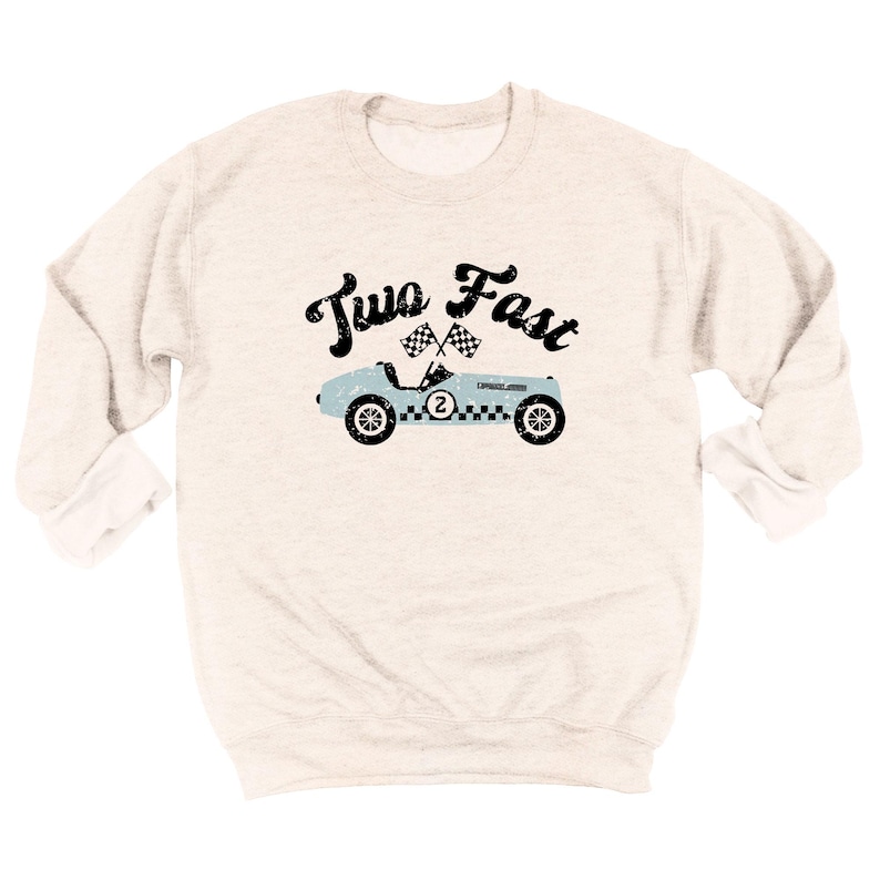 May include: A cream-colored long-sleeve sweatshirt featuring a distressed black graphic that reads Two Fast above a light blue vintage race car with a checkered flag design, the number two on the side, and two crossed checkered flags above it.