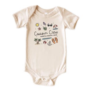 K&ouml;nnte beinhalten: Cremefarbener Baby-Body mit Strandmotiv. Das Design umfasst eine Sonne, Sonnenbrille, Palmen und den Text "Cousin Crew Myrtle Beach 2025". Der Body hat kurze &Auml;rmel und Druckkn&ouml;pfe.