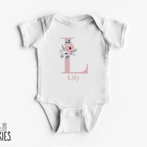 May include: White baby bodysuit with a pink floral letter "L" and the name "Lily" printed below. The bodysuit features a round neckline and short sleeves. The "little spunkies" logo is in the bottom left corner.